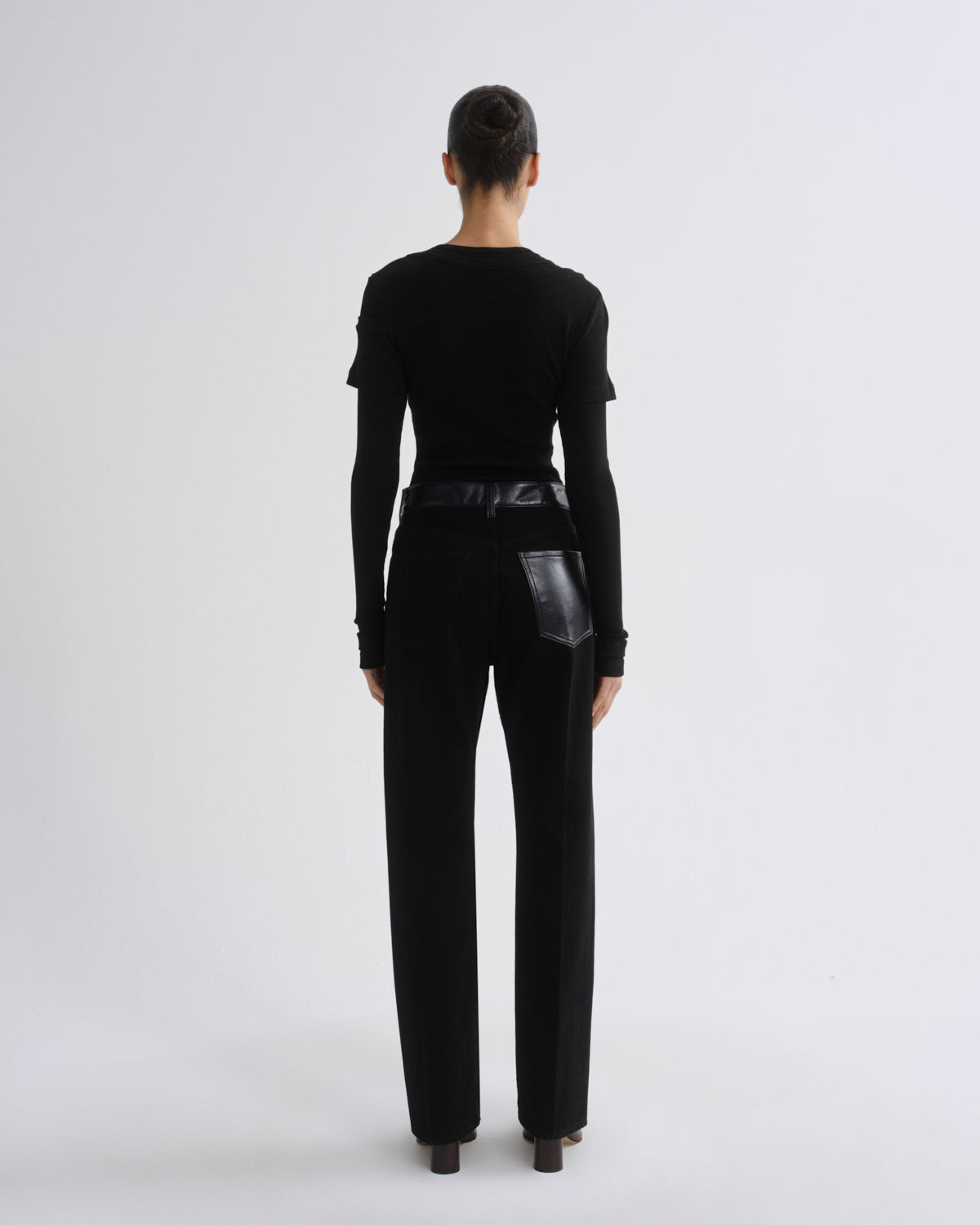 A model viewed from the back wearing a black long-sleeve top with shoulder details and black wide-leg trousers featuring a glossy black leather waist belt and a matching glossy black leather back pocket, set against a plain light gray studio background.