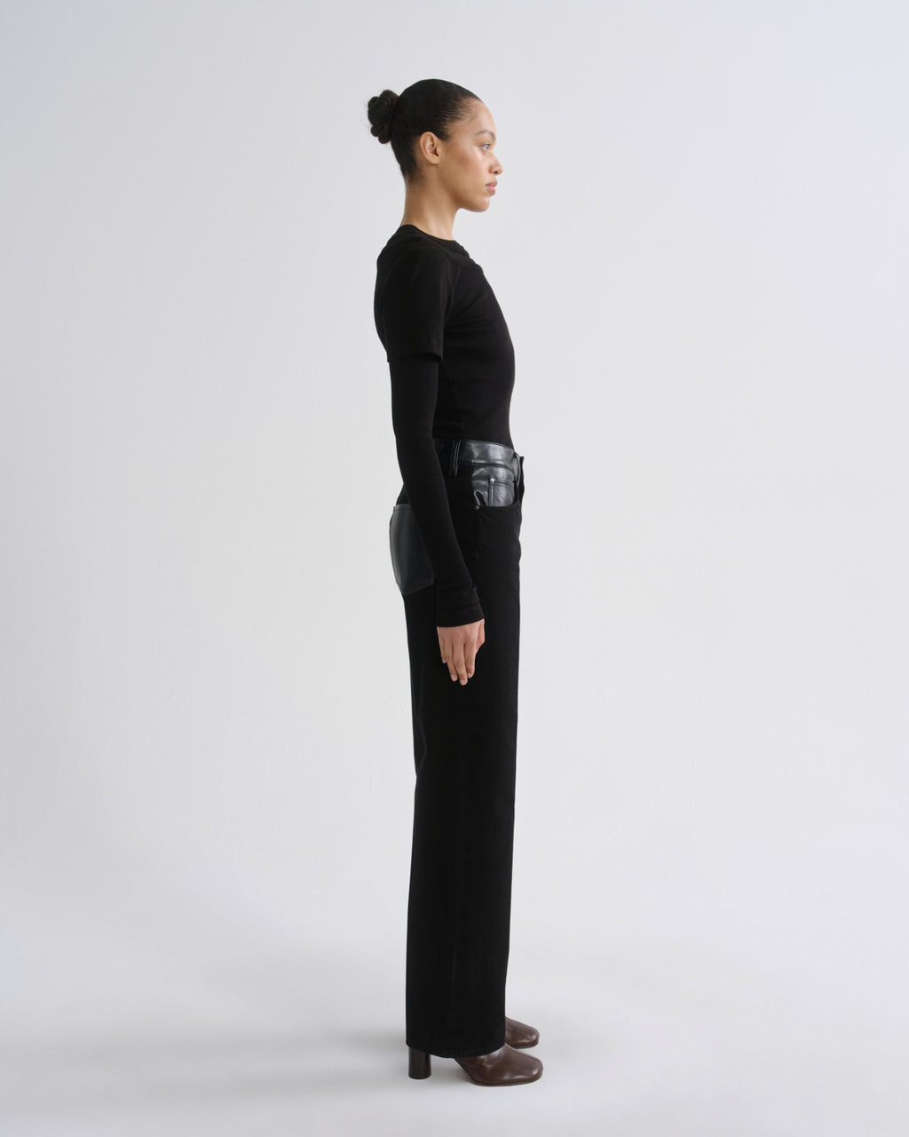 A model in profile wearing a black long-sleeve top and matching black wide-leg trousers, styled with a black belt and brown shoes, against a light gray studio background.