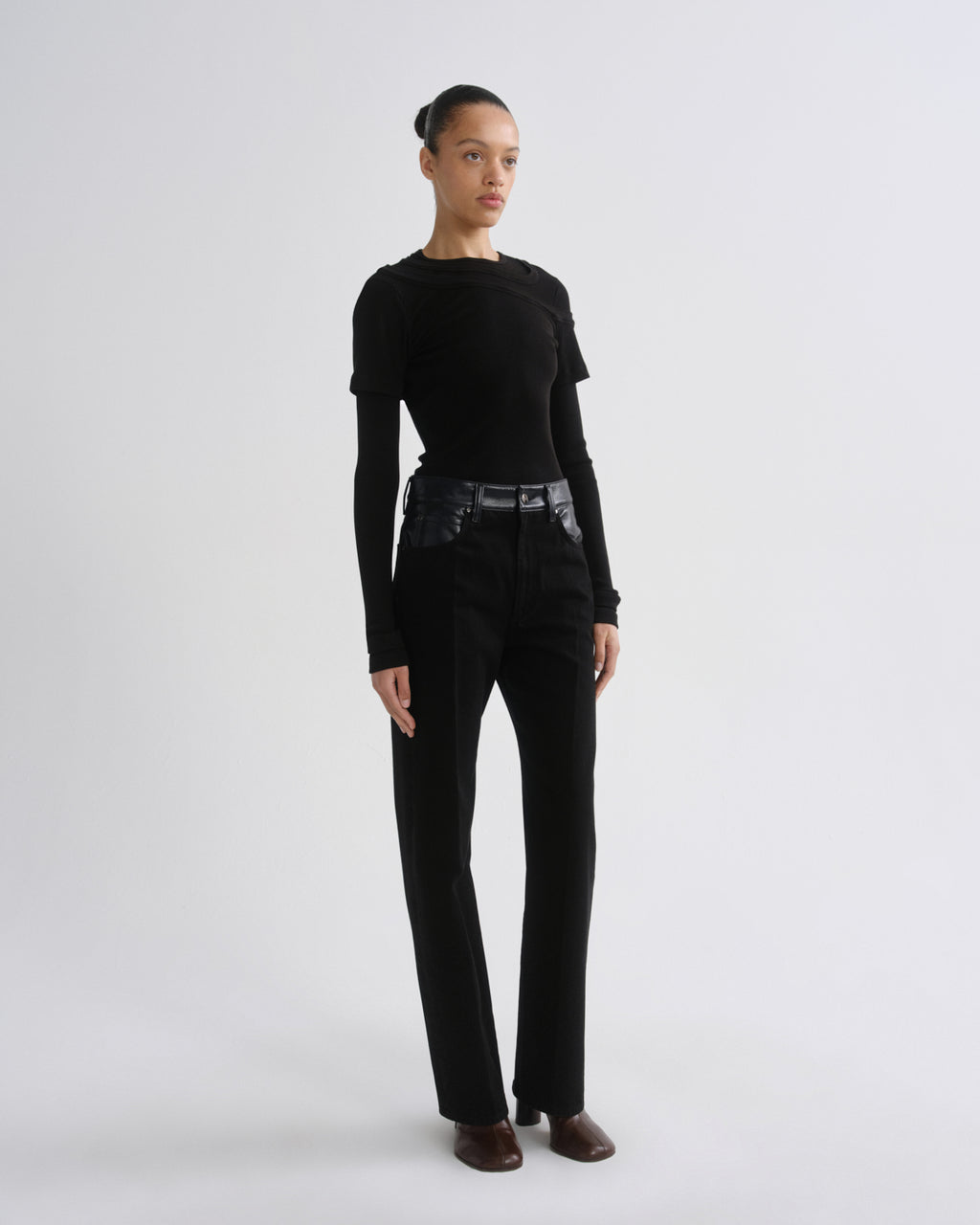A model stands in a studio wearing a black long-sleeve top, black high-waisted straight-leg pants with a wide black belt, and brown pointed shoes against a light gray background.