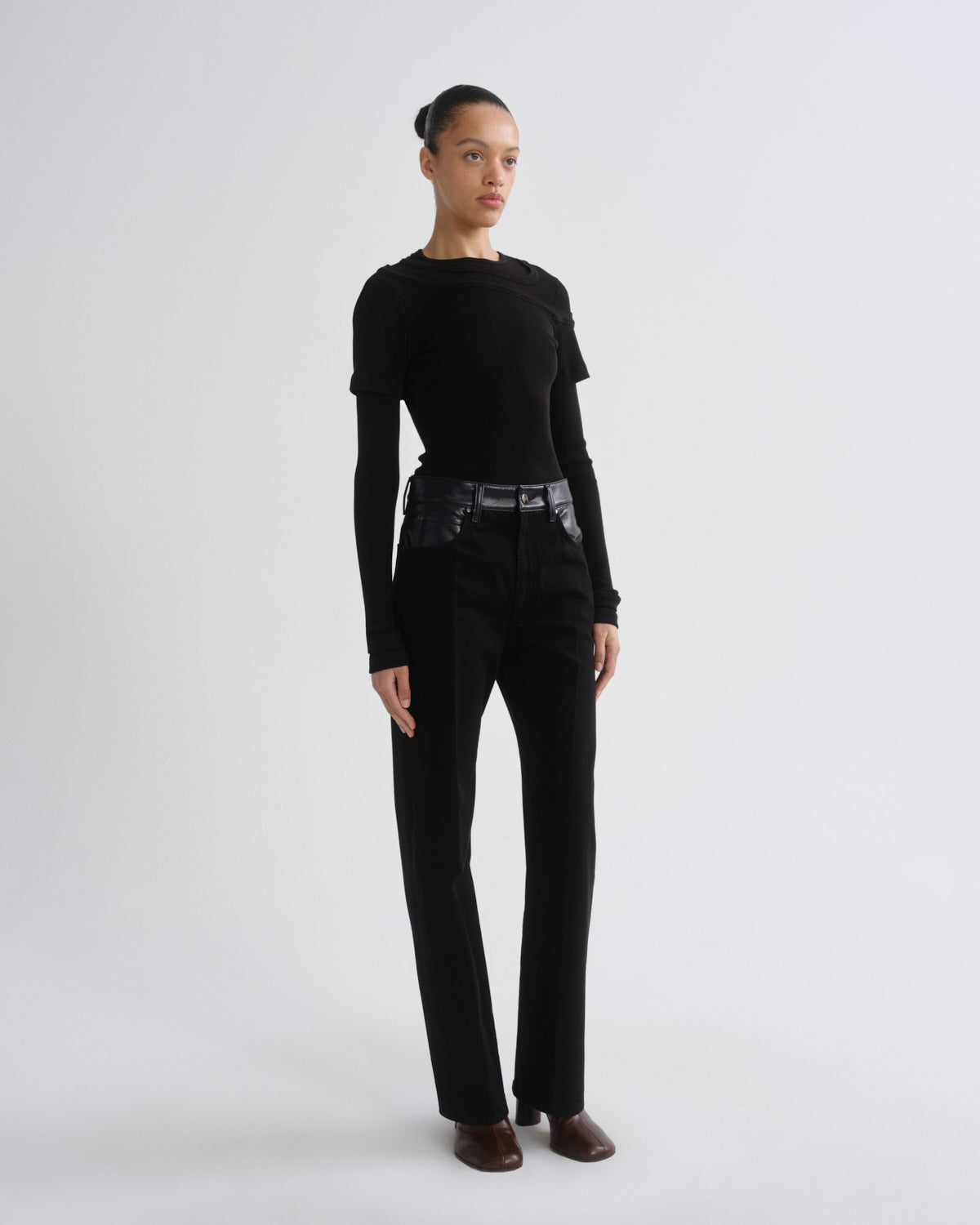 A model stands in a studio wearing a black long-sleeve top, black high-waisted straight-leg pants with a wide black belt, and brown pointed shoes against a light gray background.