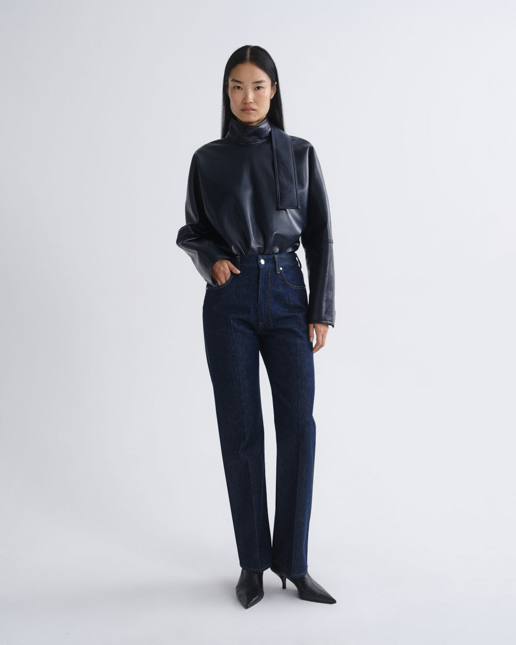 A model stands against a light gray studio background wearing a black faux-leather mock-neck jacket with structured shoulders, dark blue straight-leg jeans, and black pointed-toe heeled boots.