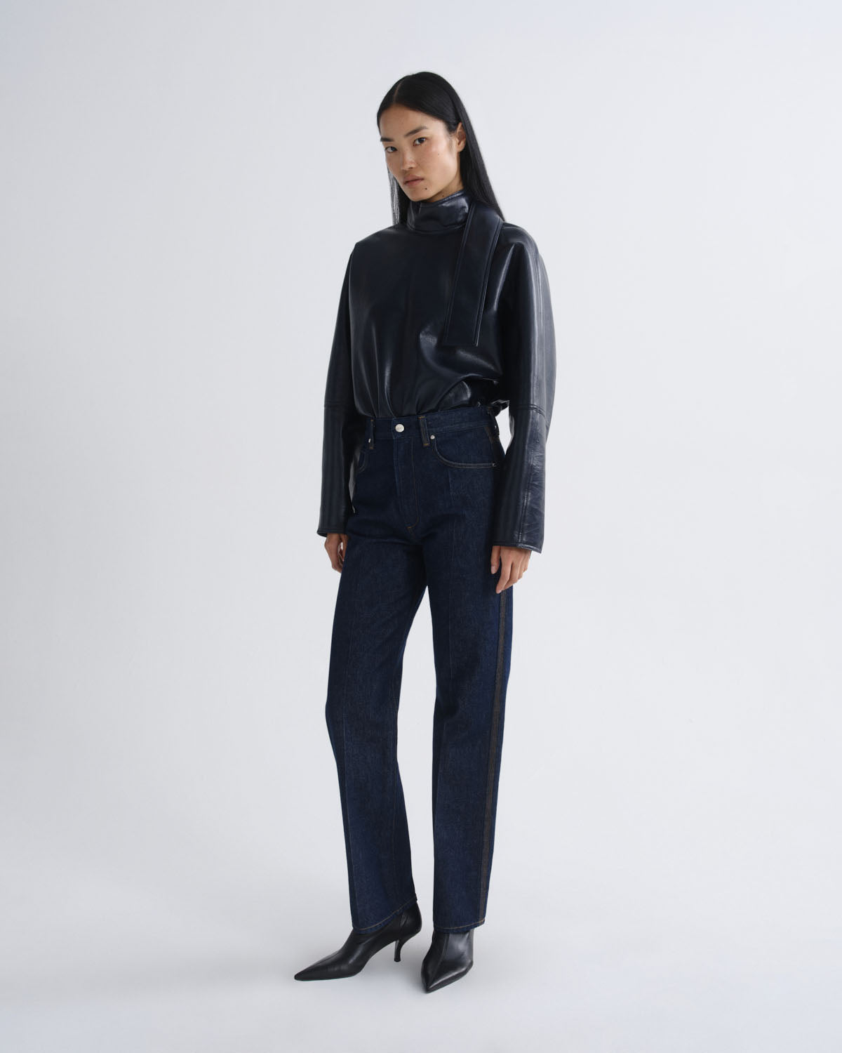A model stands against a light gray studio background wearing a black long-sleeve mock-neck top with glossy sleeves, dark blue straight-leg jeans, and black pointed-toe heeled boots.