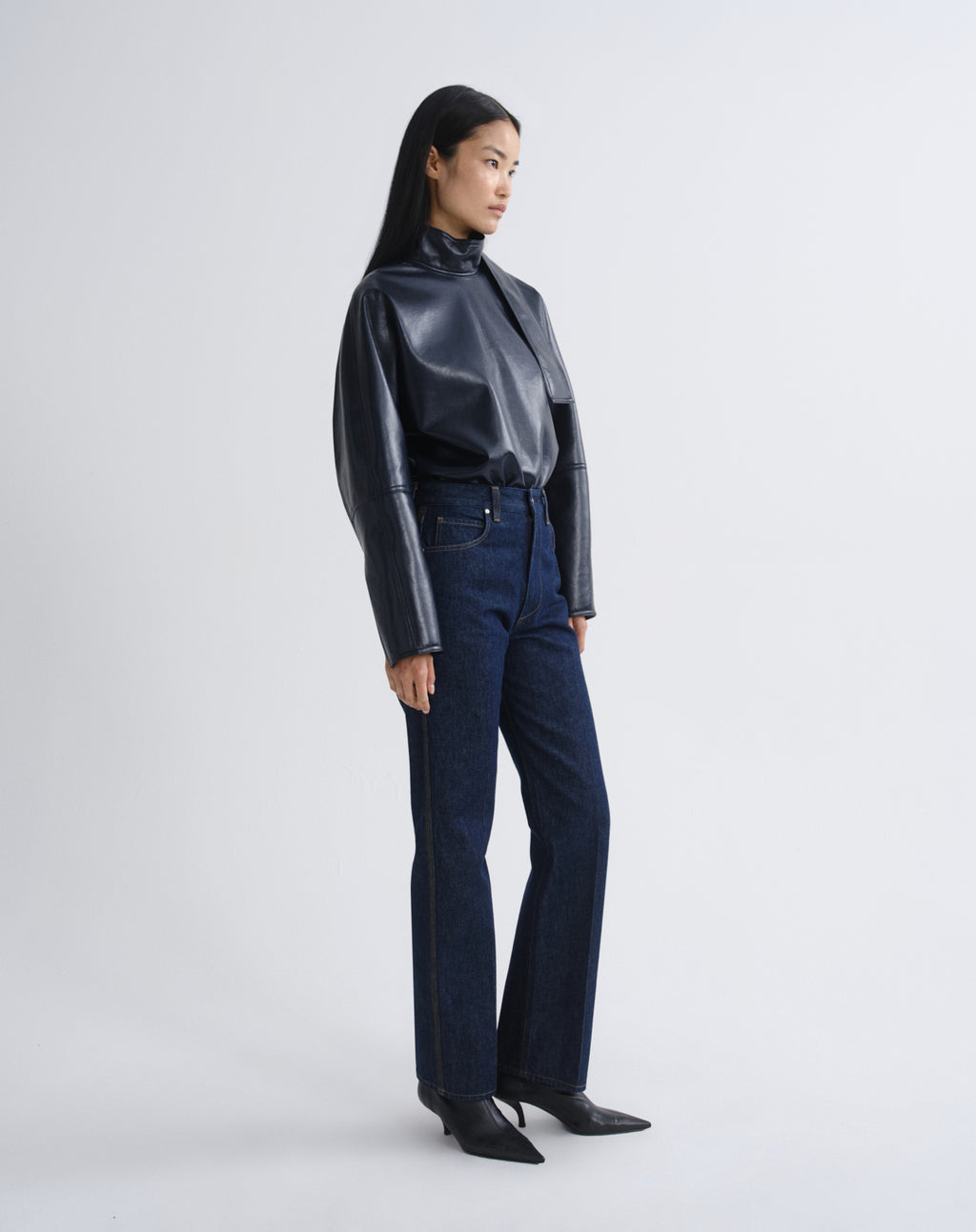 Person wearing a navy satin button-up shirt with dark indigo straight leg jeans viewed from the side on a grey background