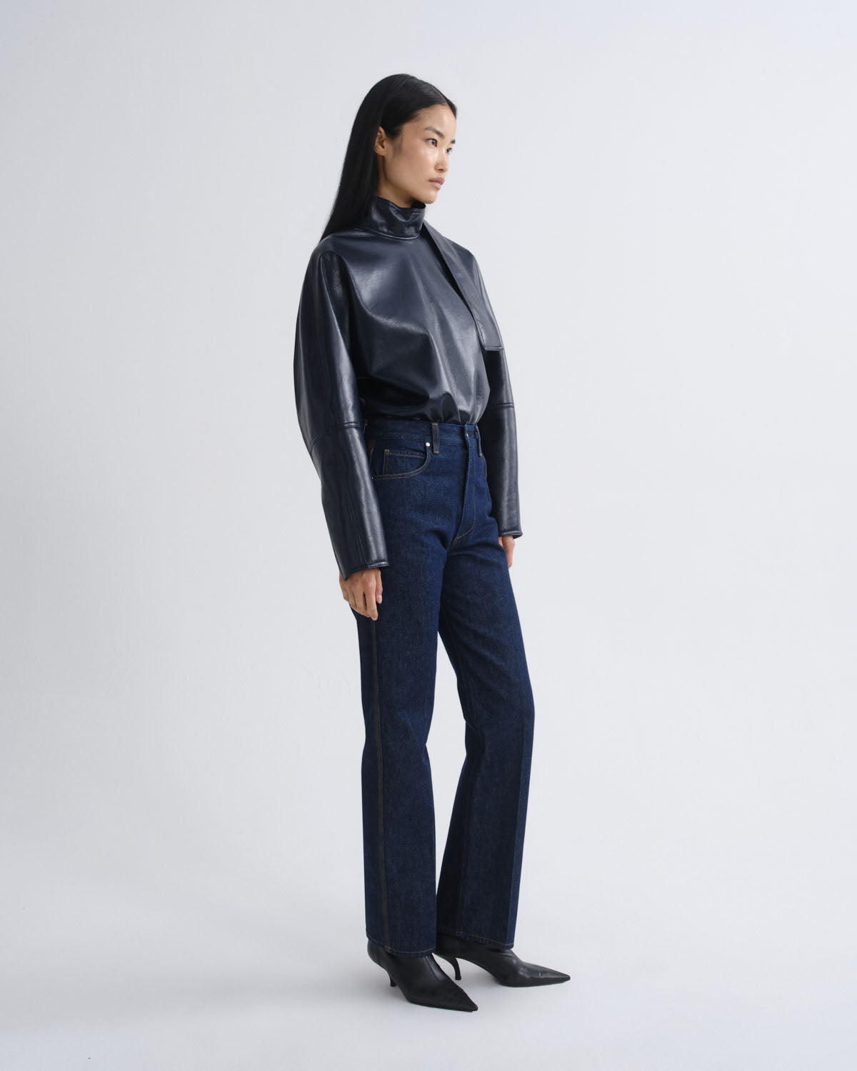 A model is shown in profile wearing a shiny black cropped long-sleeve top, dark blue high-waisted flare jeans, and black pointed-toe heels against a light gray studio background.