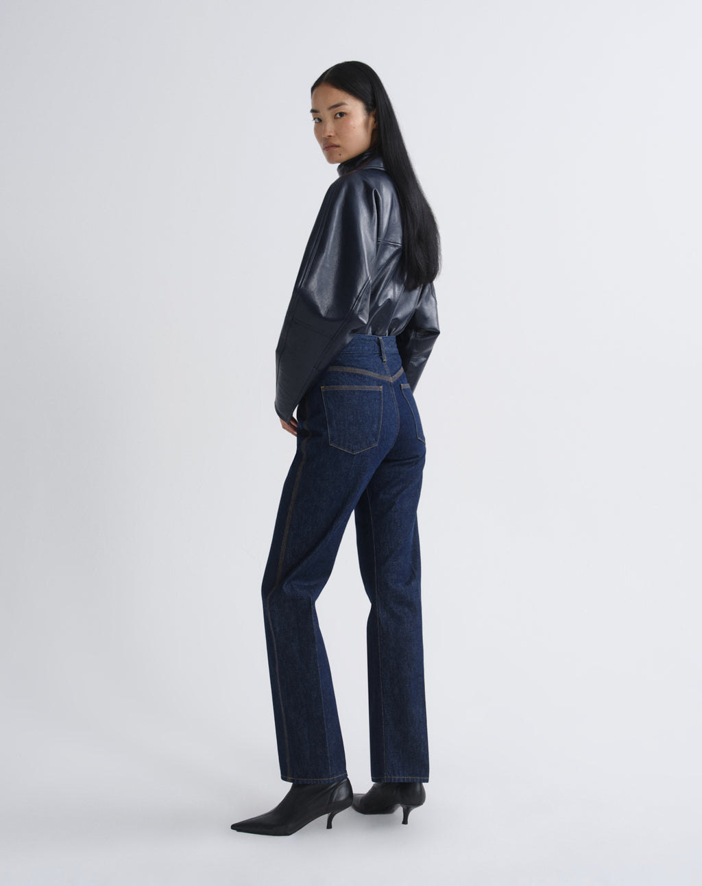 Person wearing a navy satin button-up shirt with dark indigo straight leg jeans viewed from the back on a grey background