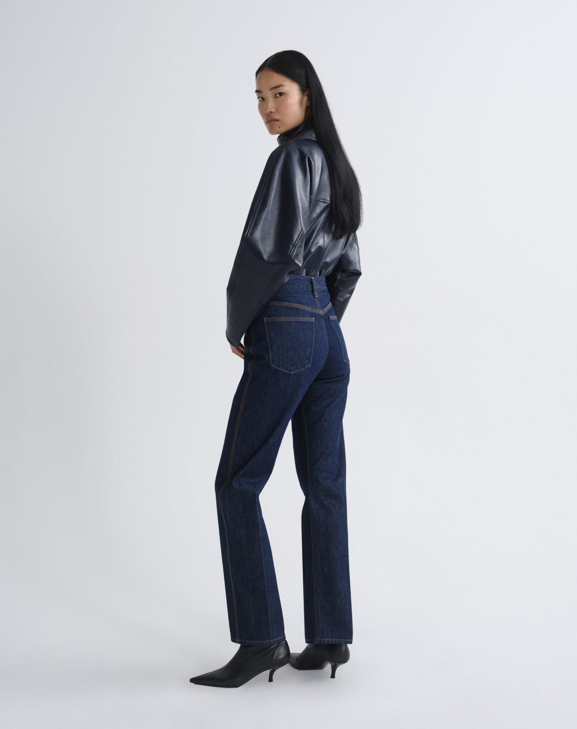 Person wearing a navy satin button-up shirt with dark indigo straight leg jeans viewed from the back on a grey background