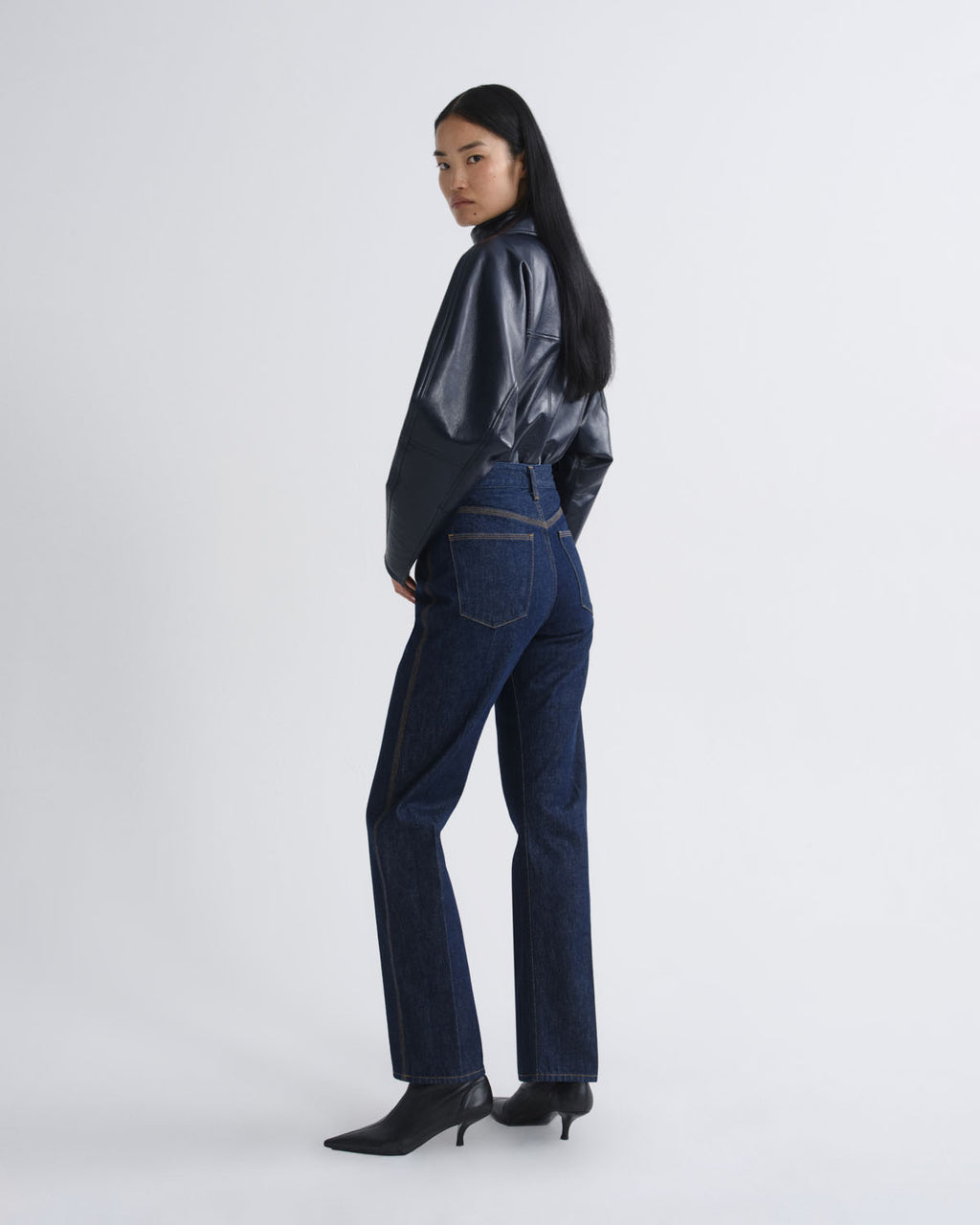 A model is shown wearing a shiny black cropped long-sleeve top, dark blue high-waisted flare jeans, and black pointed-toe heels against a light gray studio background.