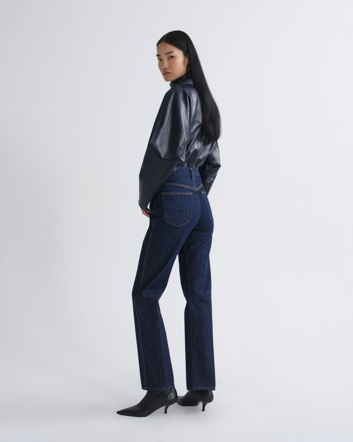 A model is shown wearing a shiny black cropped long-sleeve top, dark blue high-waisted flare jeans, and black pointed-toe heels against a light gray studio background.