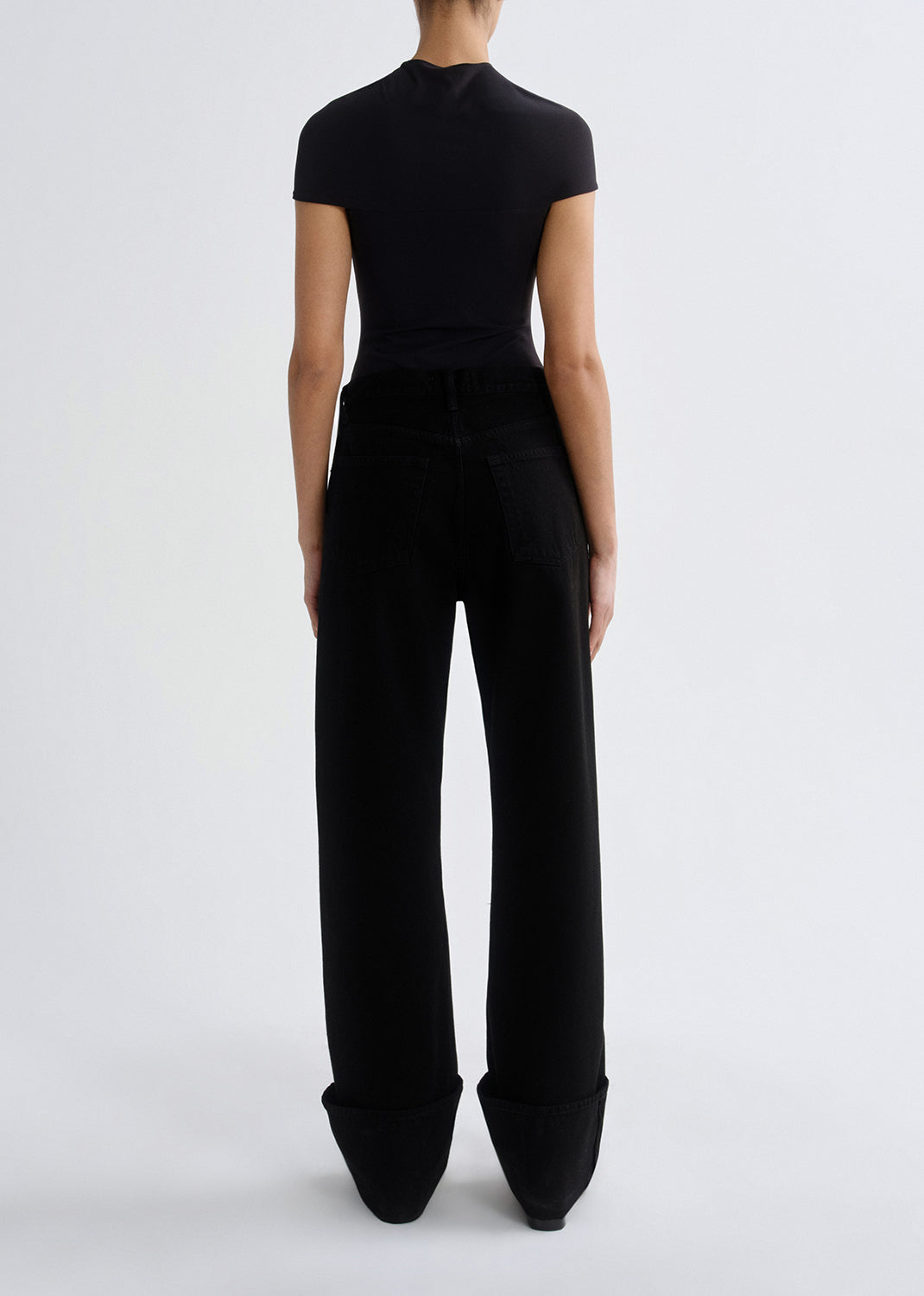 Person wearing a black cap sleeve draped top with black straight leg jeans viewed from the back on a grey background