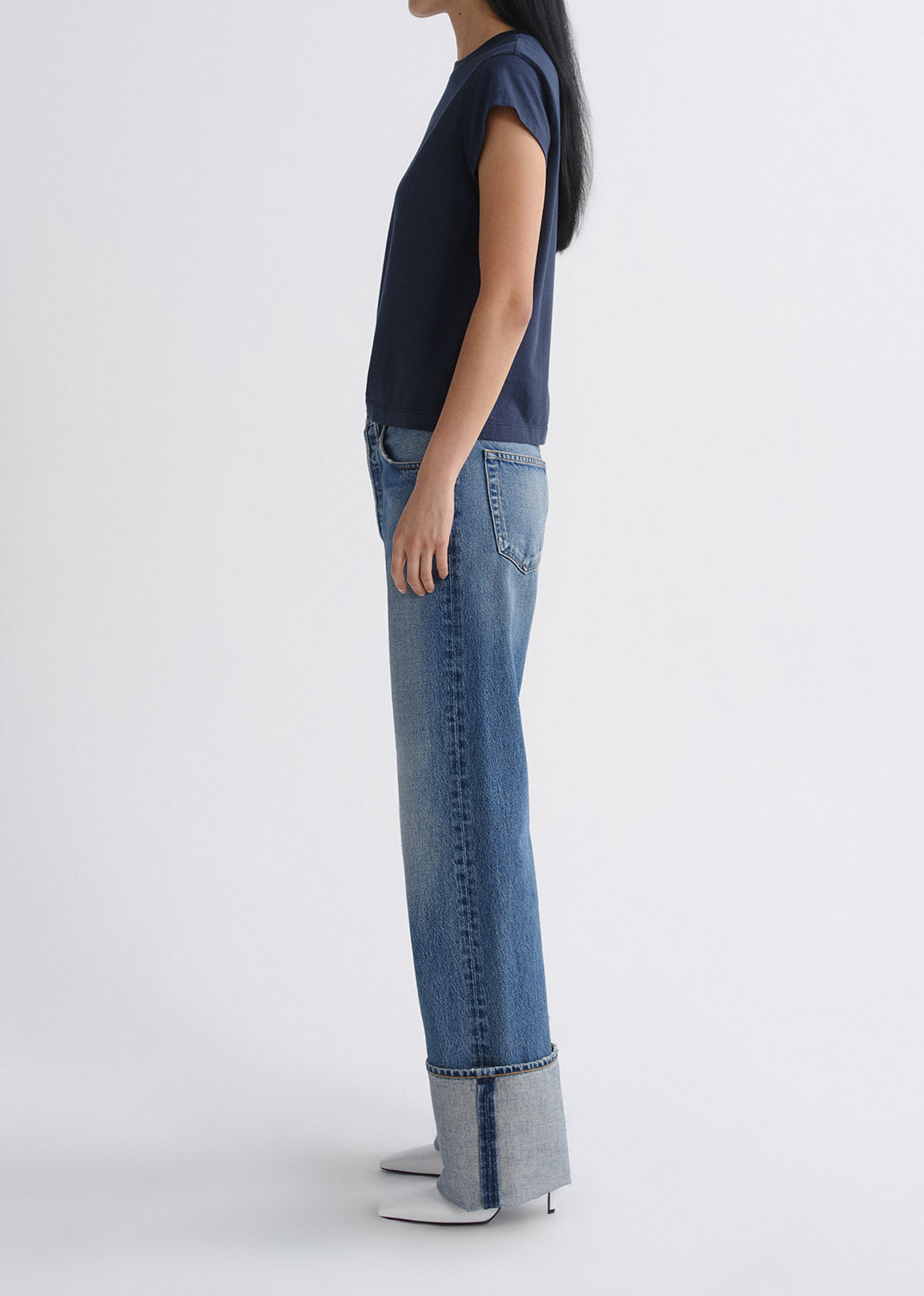 Person wearing a navy cap sleeve top with light wash cuffed straight leg jeans viewed from the side on a grey background.