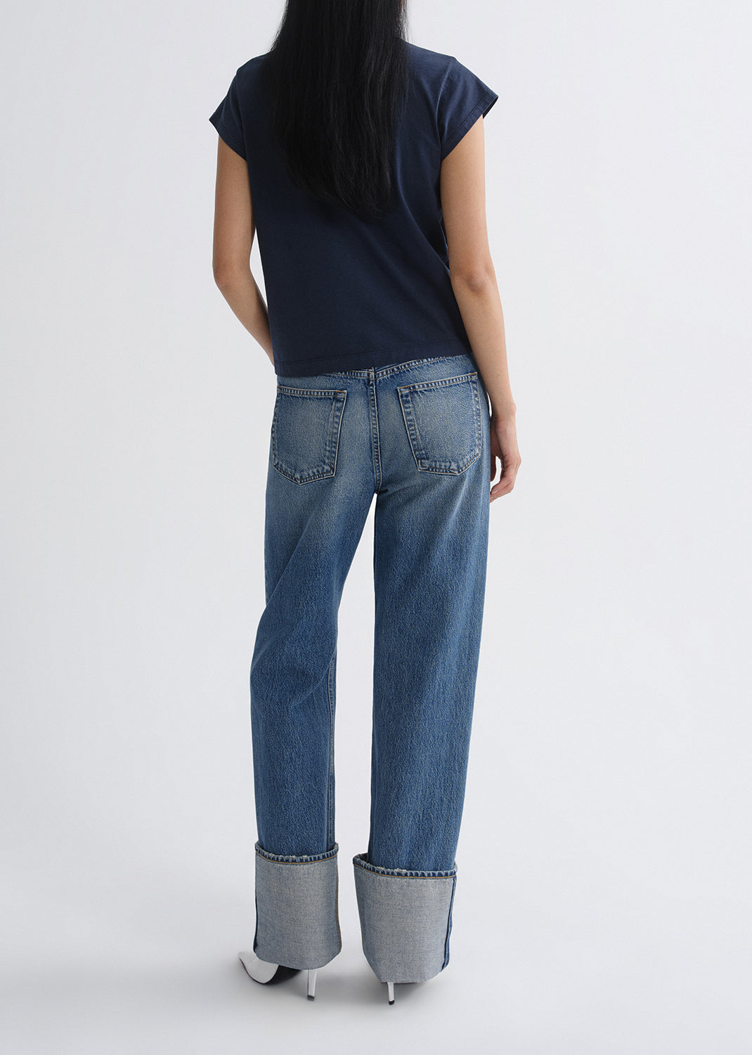 Person wearing a navy cap sleeve top with light wash cuffed straight leg jeans viewed from the back on a grey background.