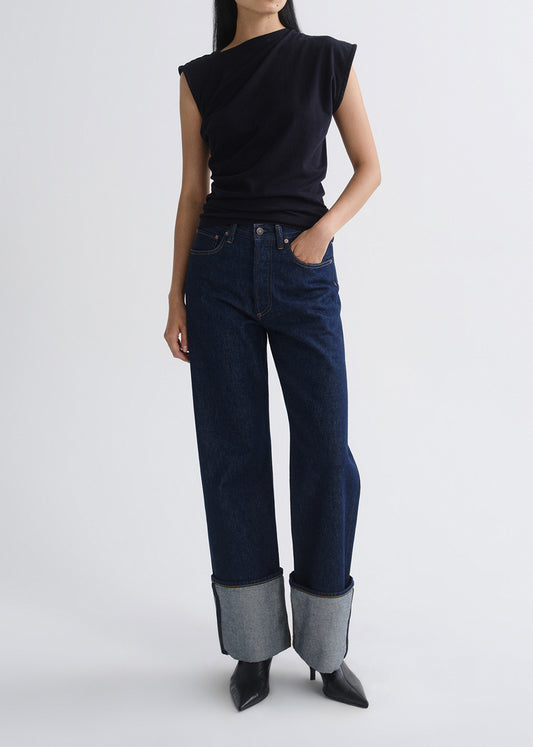 A model is shown from the neck down wearing a black asymmetrical sleeveless top, dark blue wide-leg jeans with rolled cuffs, and black pointed-toe heels against a grey background.