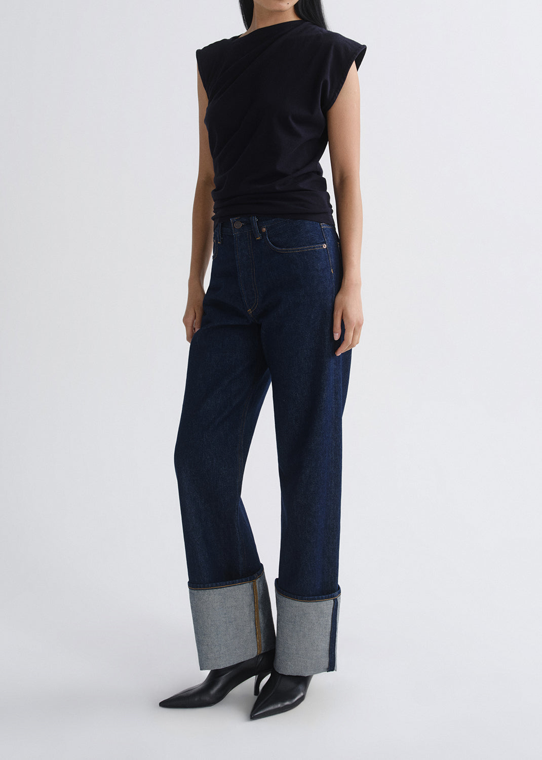 A model is shown from the back wearing dark blue wide-leg jeans with a rolled gray hem, a black sleeveless top, and black pointed-toe heels against a grey background.