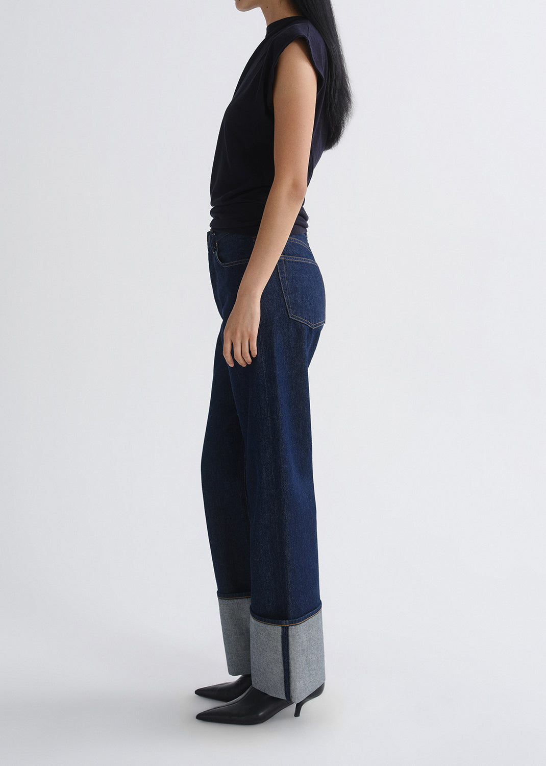 A model shown in profile wearing dark blue wide-leg denim jeans with light gray turned-up cuffs, a black sleeveless top, and black pointed-toe heels, against a grey background.