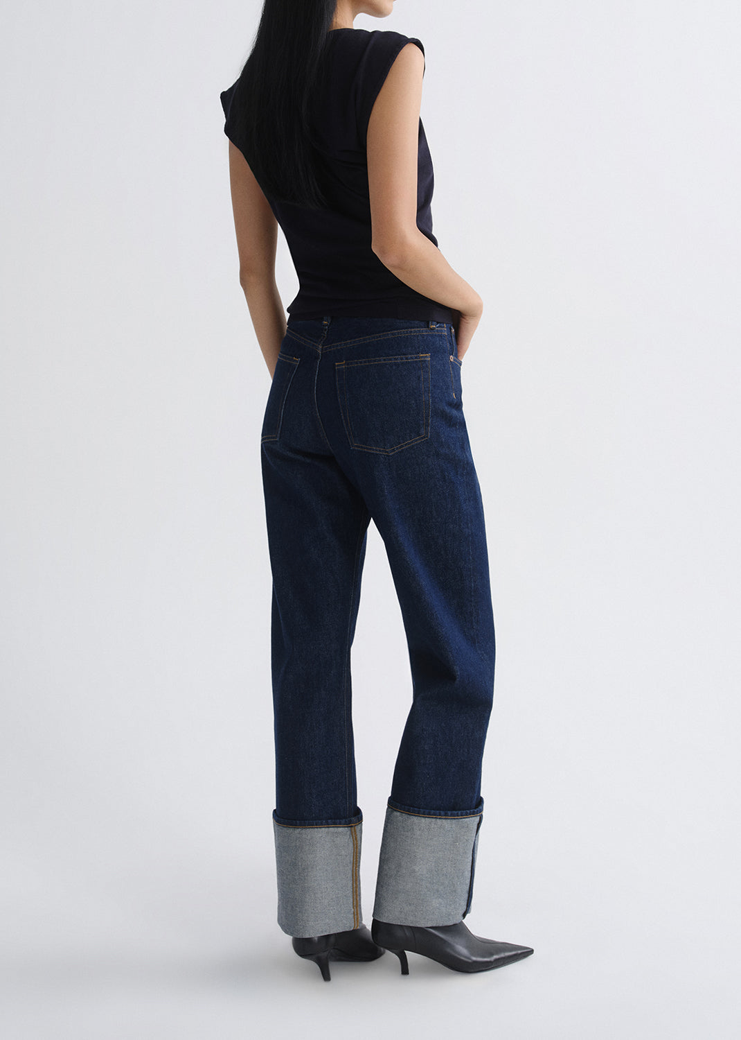 A model shown from the back wearing dark blue cropped jeans with rolled cuffs, a black sleeveless top, and black pointed-toe heels against a grey background.
