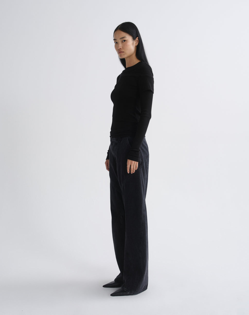 A model stands in profile wearing a black long-sleeve top and black wide-leg trousers against a plain light grey background.