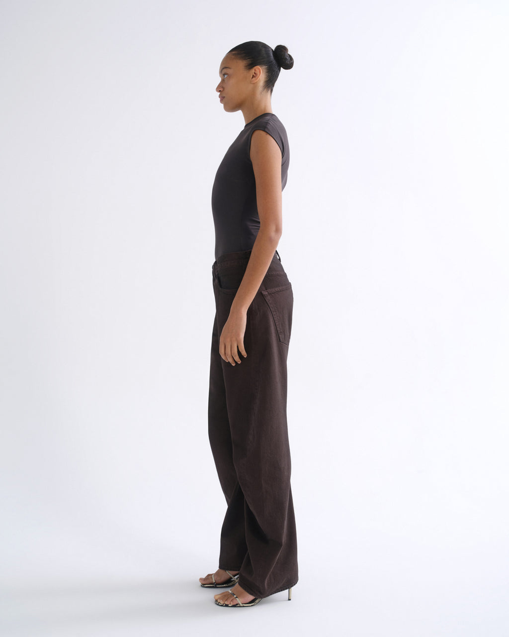 A model in side profile wearing a dark brown sleeveless top and matching wide-leg trousers, standing against a clean white studio background.