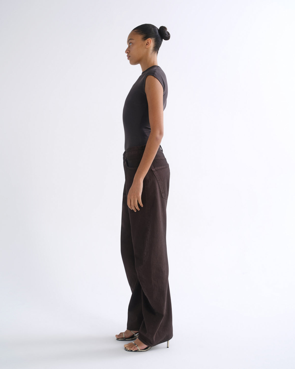 A model in side profile wearing a dark brown sleeveless top and matching wide-leg trousers, standing against a clean white studio background.