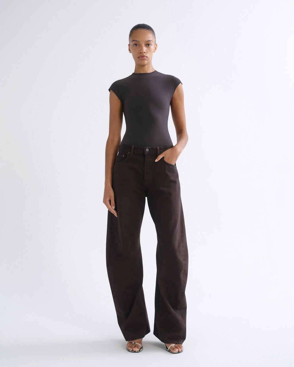 A model stands against a light gray studio background wearing a dark charcoal gray short-sleeve mock-neck top and dark brown wide-leg trousers.