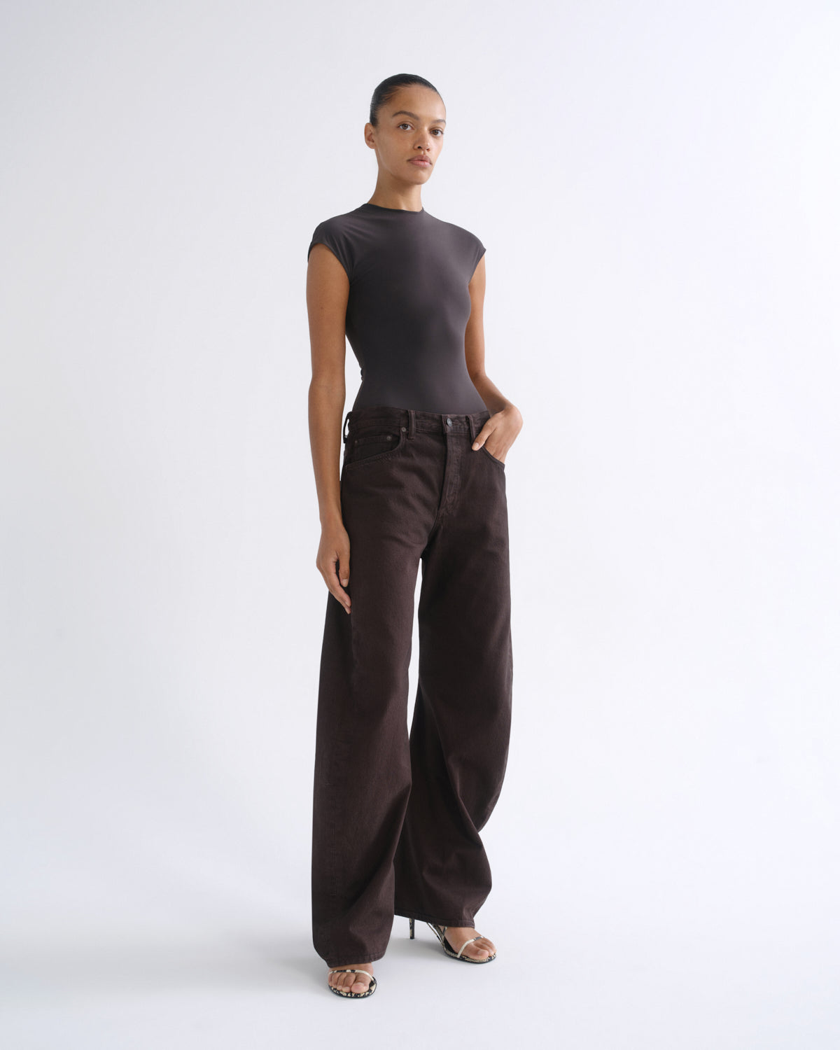 A model stands in a studio wearing a dark brown sleeveless mock-neck top and matching dark brown wide-leg trousers, paired with metallic sandals, against a light gray background.
