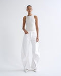 A model stands against a plain light gray studio background wearing a fitted sleeveless white top and very wide-leg white pants, with white pointed shoes.