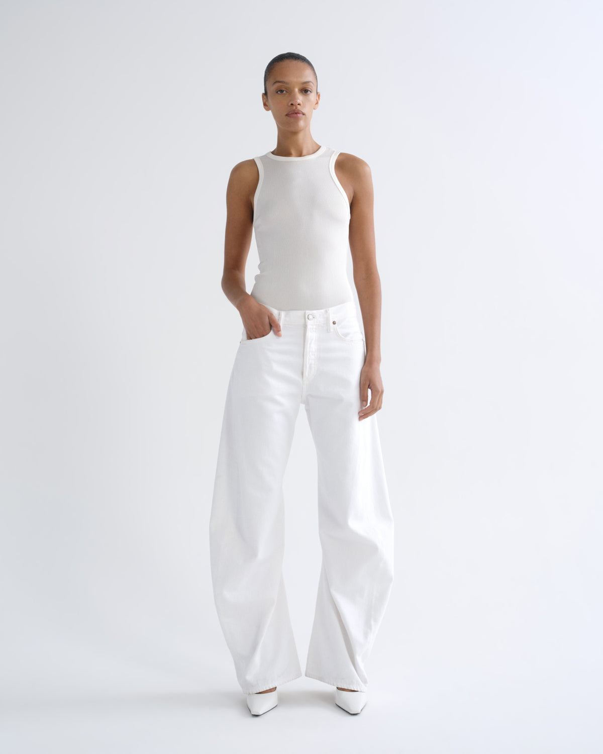 A model stands against a plain light gray studio background wearing a fitted sleeveless white top and very wide-leg white pants, with white pointed shoes.