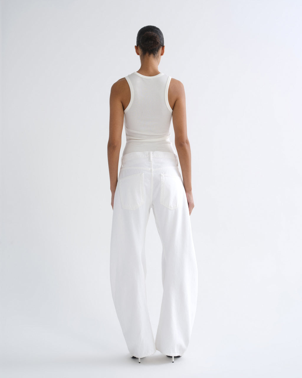 A model viewed from the back wearing a white ribbed tank top and very wide-leg white trousers against a clean white studio background.