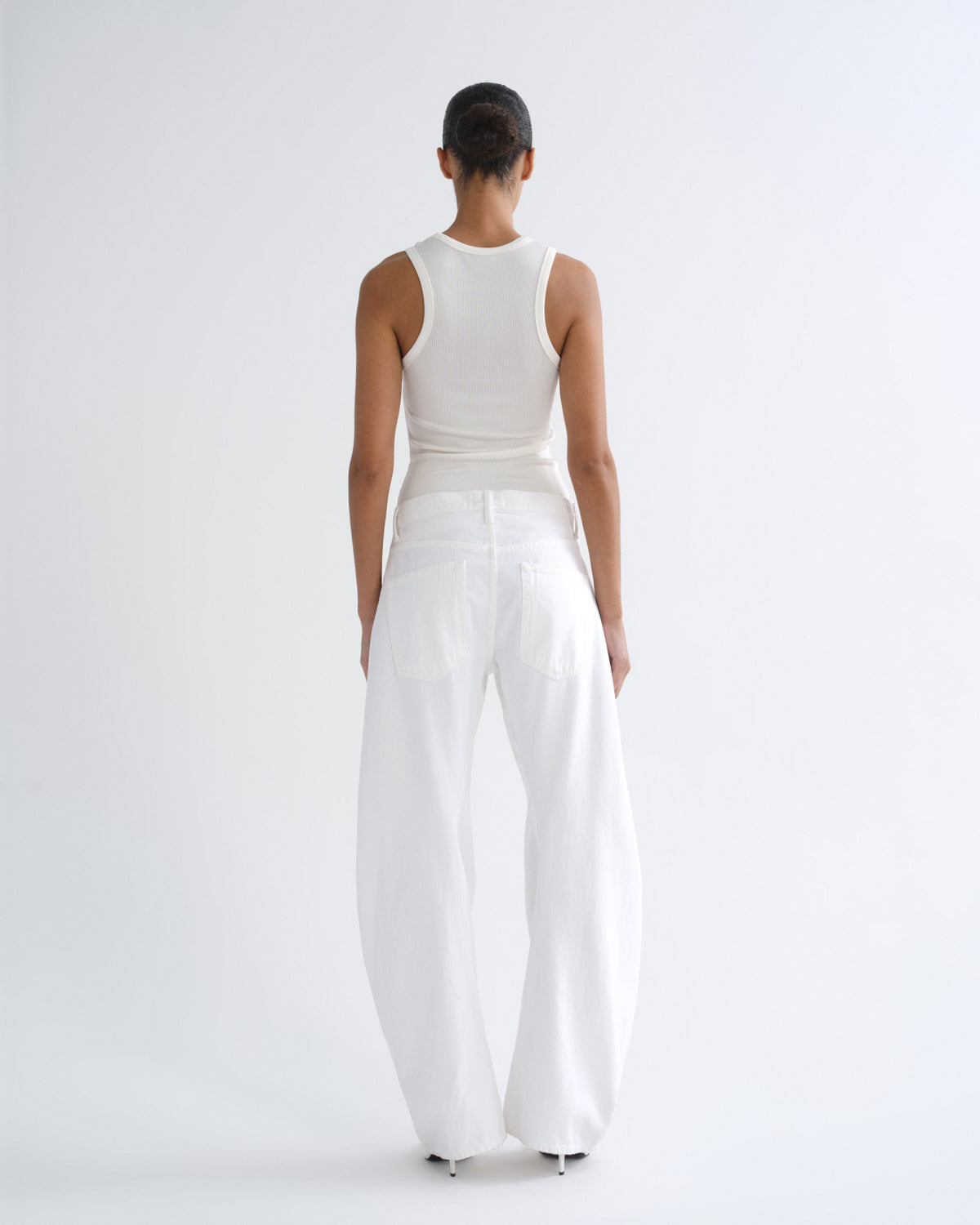 A model viewed from the back wearing a white ribbed tank top and very wide-leg white trousers against a clean white studio background.