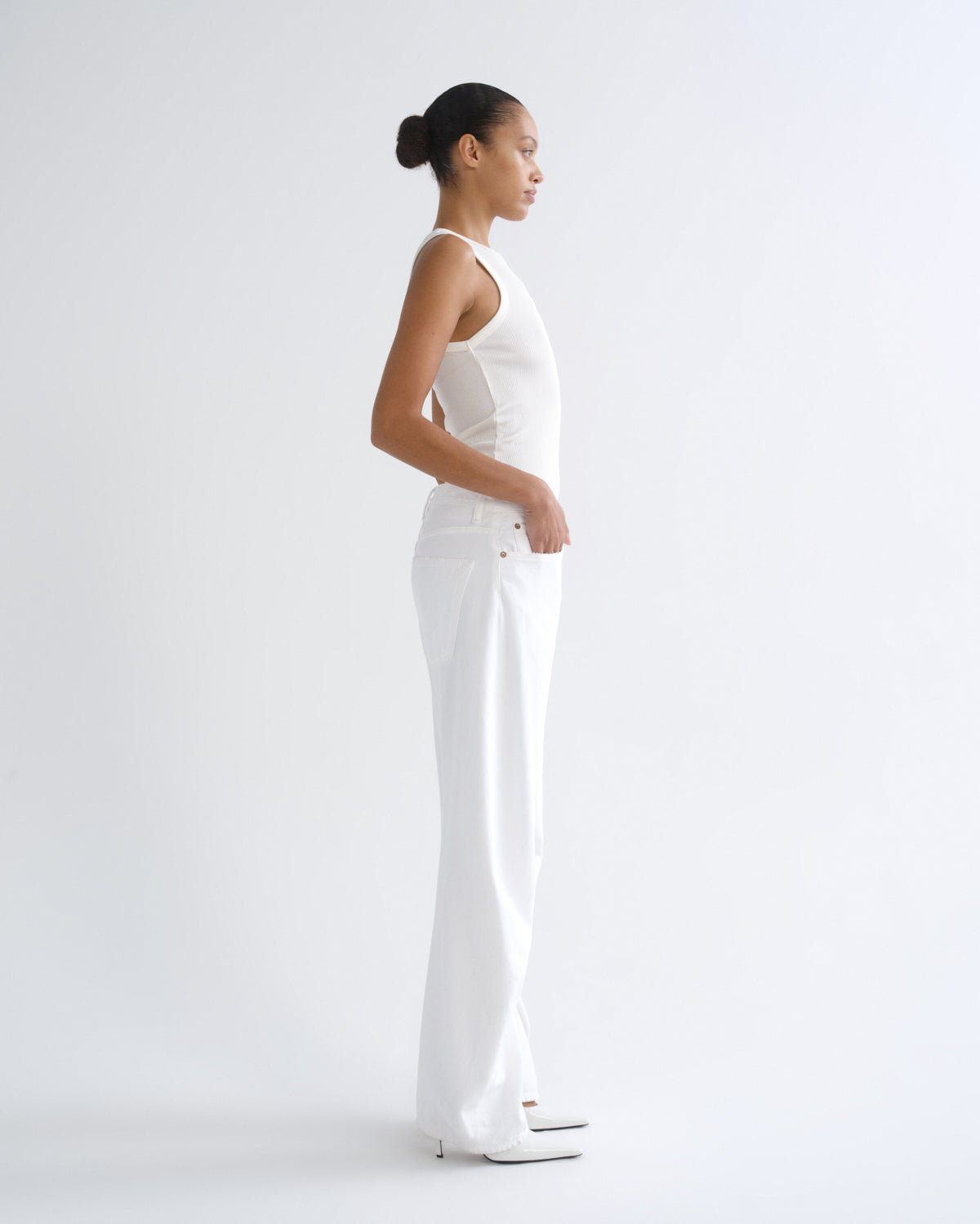 A woman in a white sleeveless top and white wide-leg pants stands in profile against a clean white studio background.