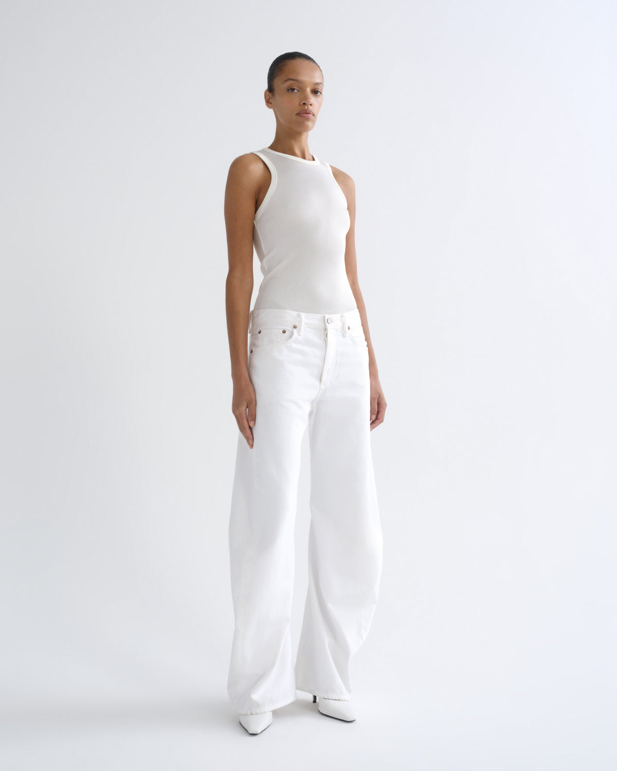 A woman in a white sleeveless top and white wide-leg pants stands in side profile against a clean white studio background.