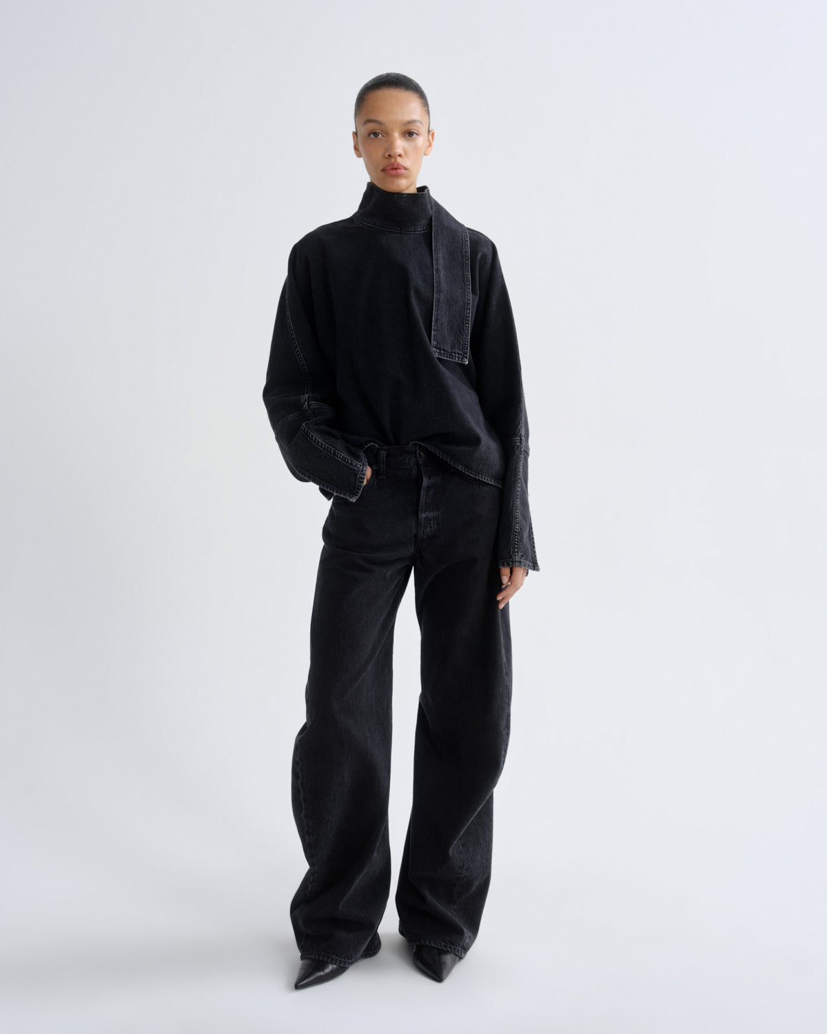 A model stands against a light gray studio background wearing a black high-neck long-sleeve top with contrasting dark denim paneling and matching black wide-leg pants.