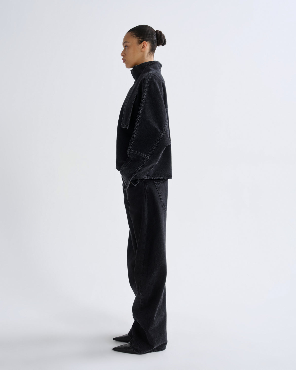 A model in profile wearing a black cropped jacket and matching black wide-leg jeans against a plain light gray studio background.