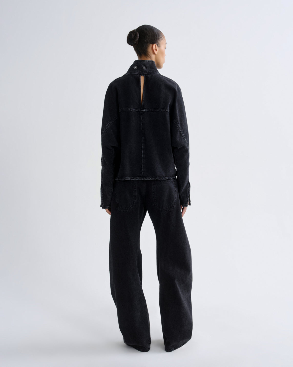 A model viewed from the back wearing a black denim long-sleeve top with a high collar and a small back slit, paired with matching wide-leg black denim pants, against a light gray studio background.