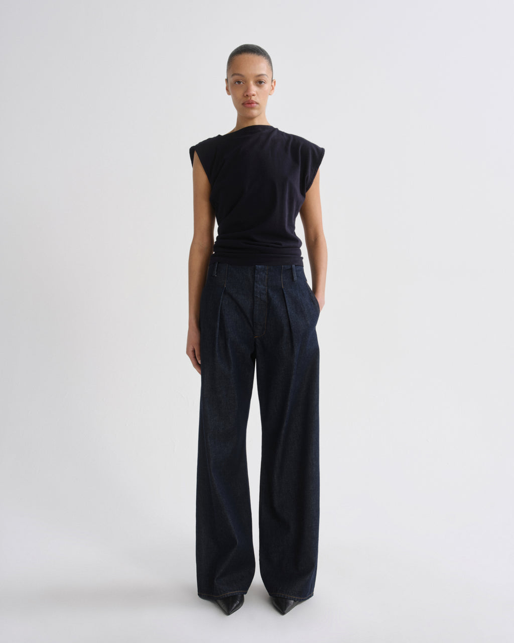 A model standing against a light gray studio background, wearing a black sleeveless top and dark wide-leg pleated trousers.