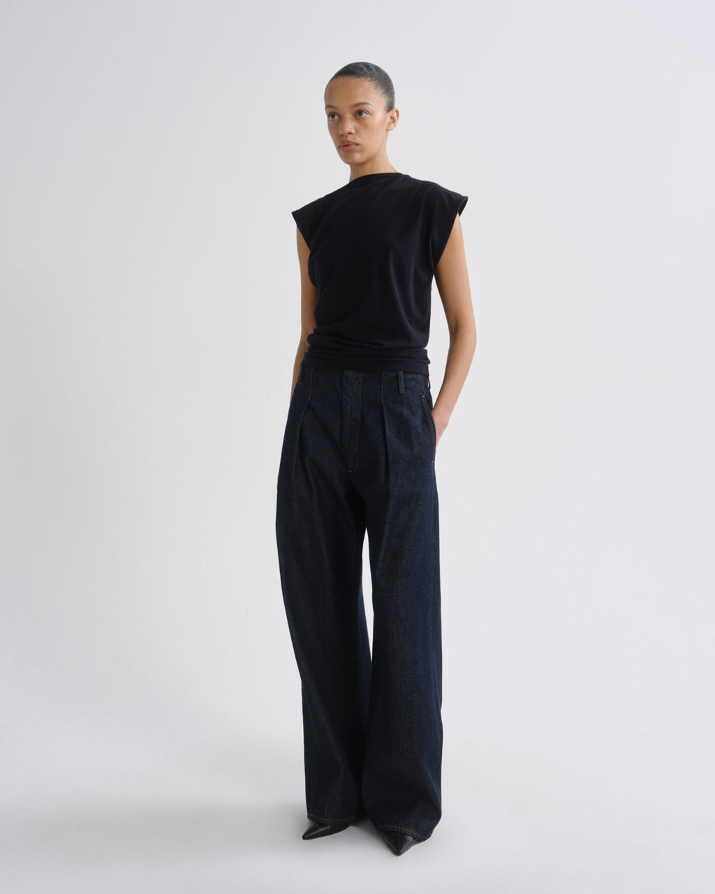 A model stands in a studio wearing a black sleeveless top and dark wide-leg trousers, photographed against a plain light gray background.