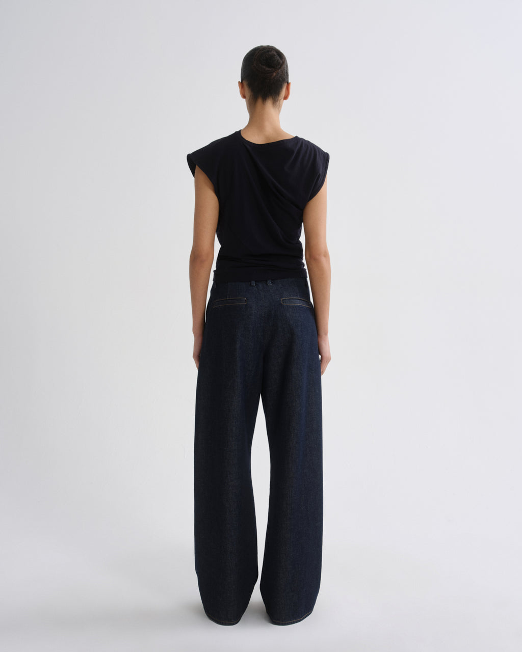 A model shown from the back wearing a black draped sleeveless top and dark wide-leg denim pants against a light gray studio background.