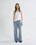 A model stands against a light gray studio background wearing a white ribbed tank top, light-wash wide-leg pleated jeans, and black pointed shoes.