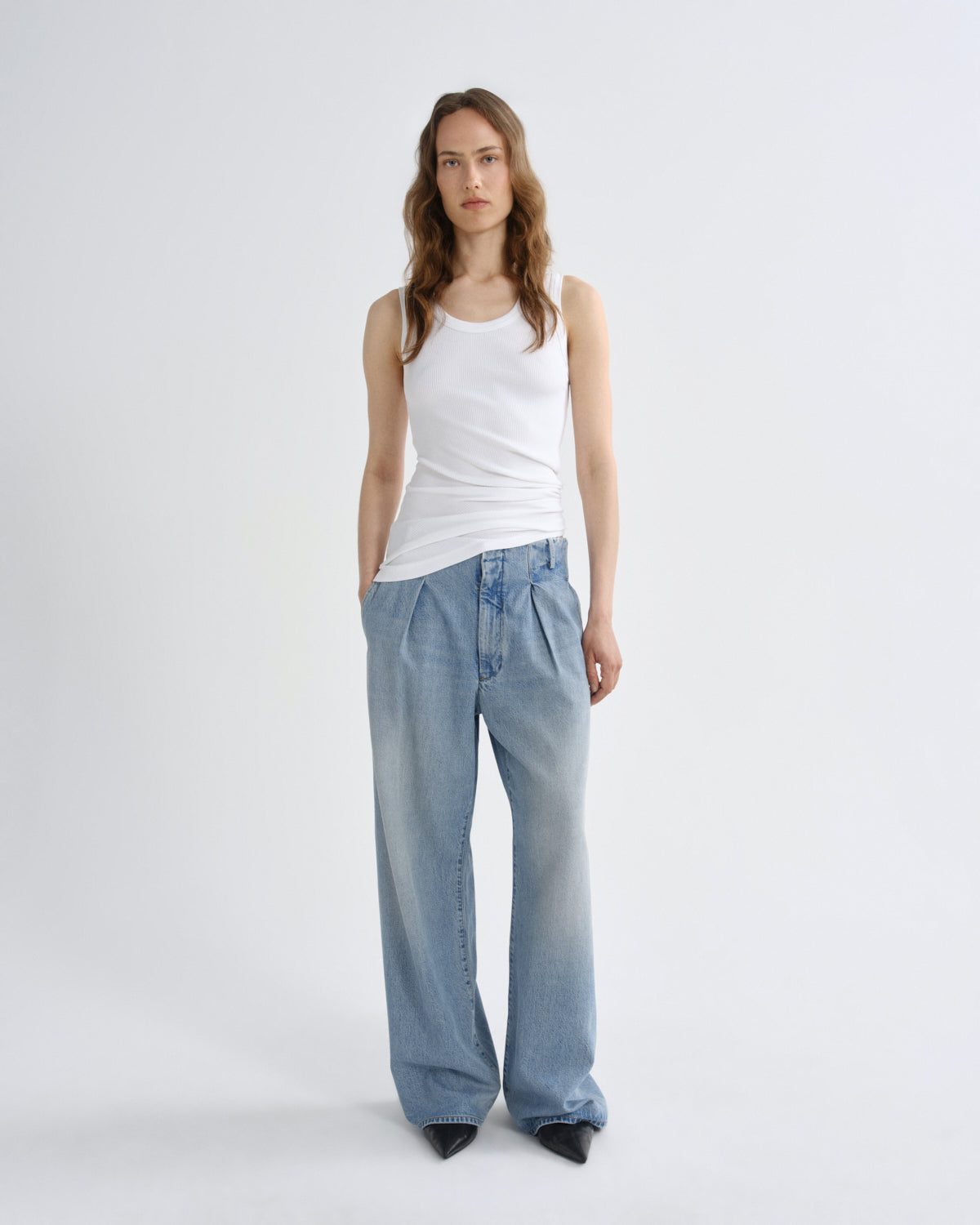 A model stands against a light gray studio background wearing a white ribbed tank top, light-wash wide-leg pleated jeans, and black pointed shoes.