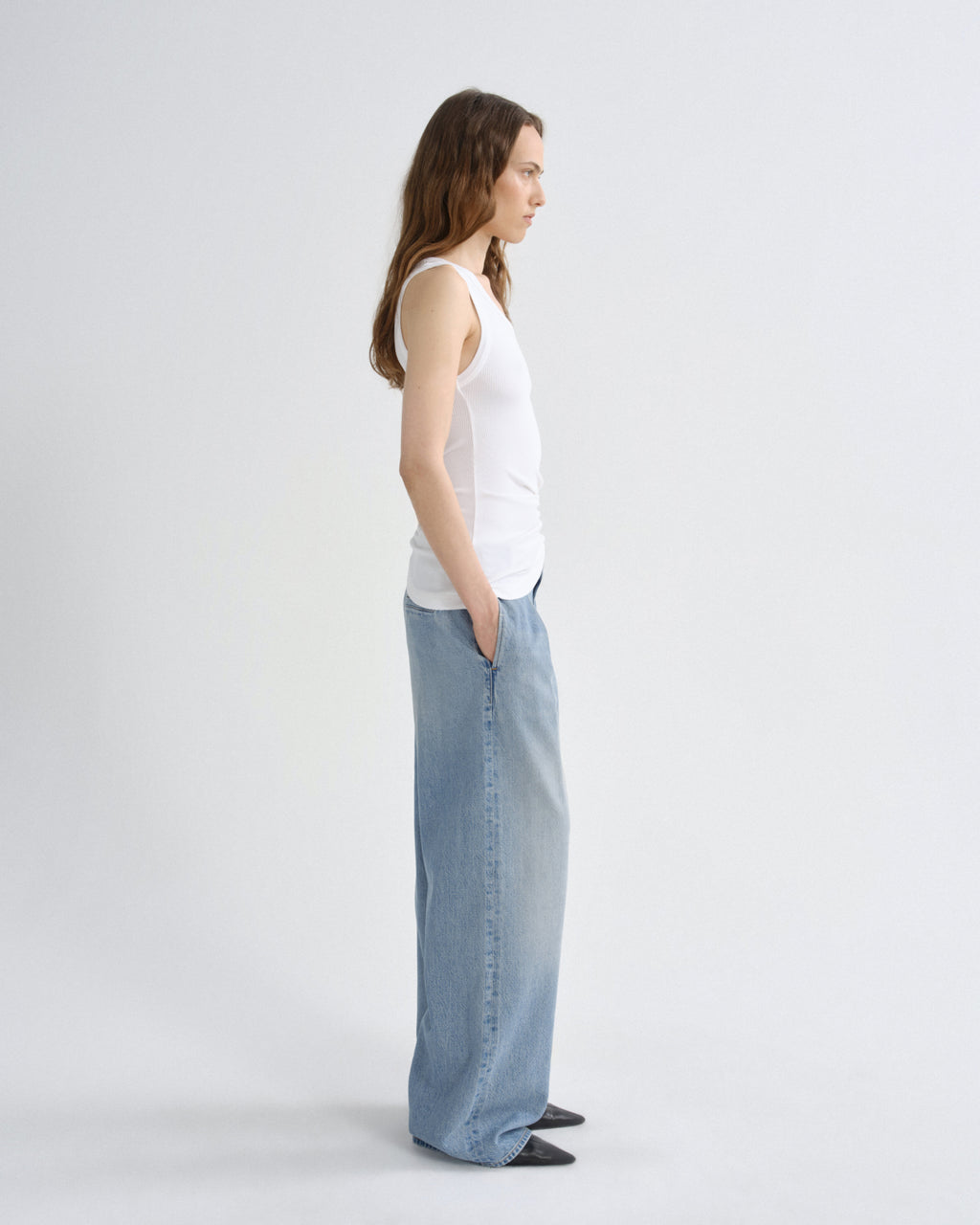 A woman stands in profile wearing a white ribbed tank top and light-wash wide-leg denim jeans against a plain light gray studio background.