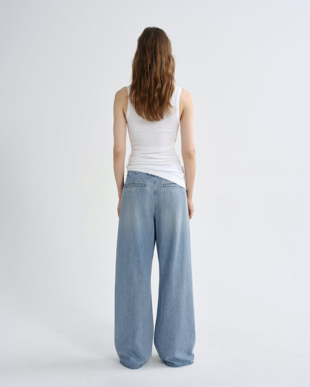 A woman viewed from behind wearing a white ribbed tank top and light-wash wide-leg denim jeans against a plain light gray studio background.