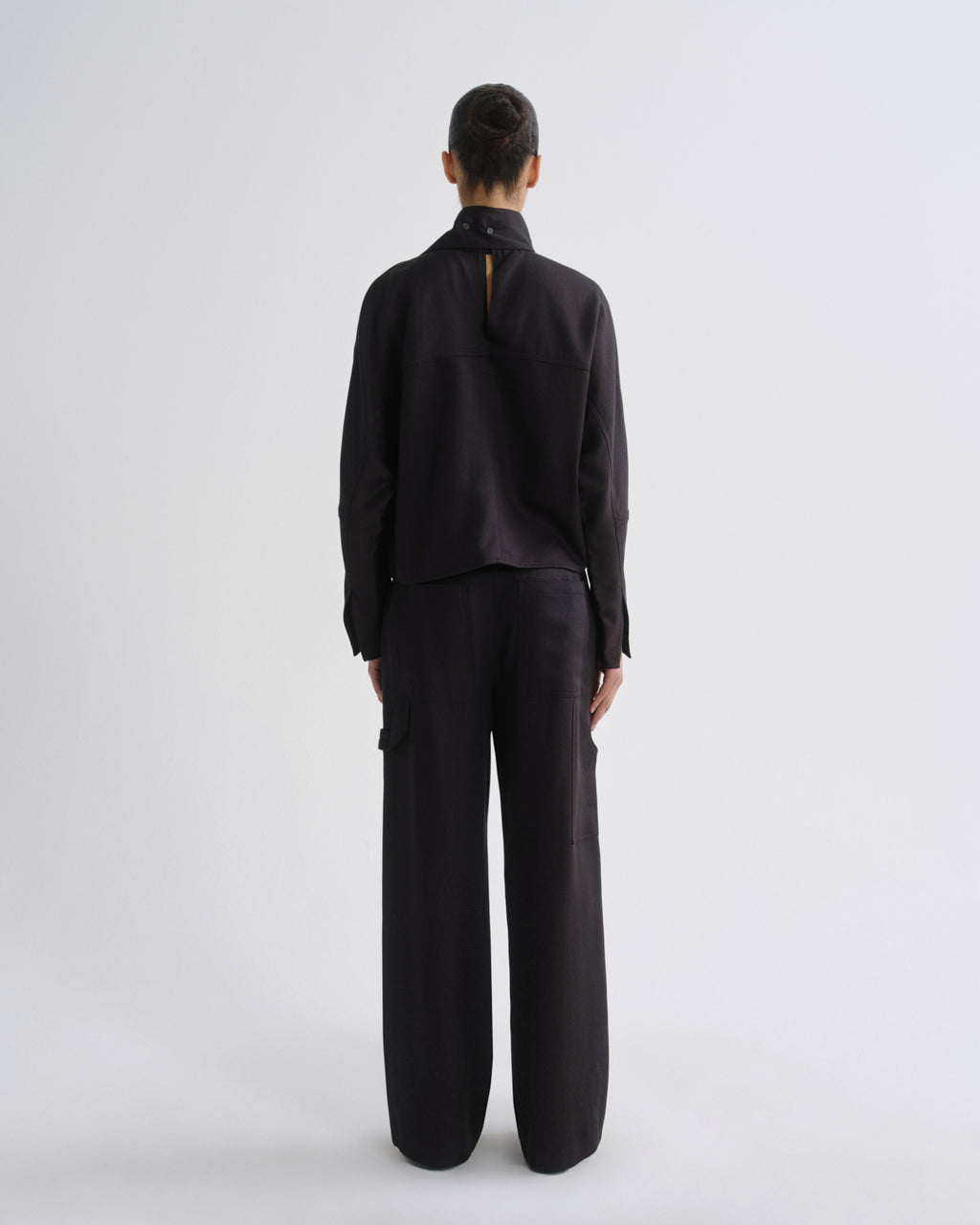 A model viewed from the back wearing a charcoal long-sleeve top with a high mock neck and wide-leg charcoal trousers, set against a plain light gray studio background.