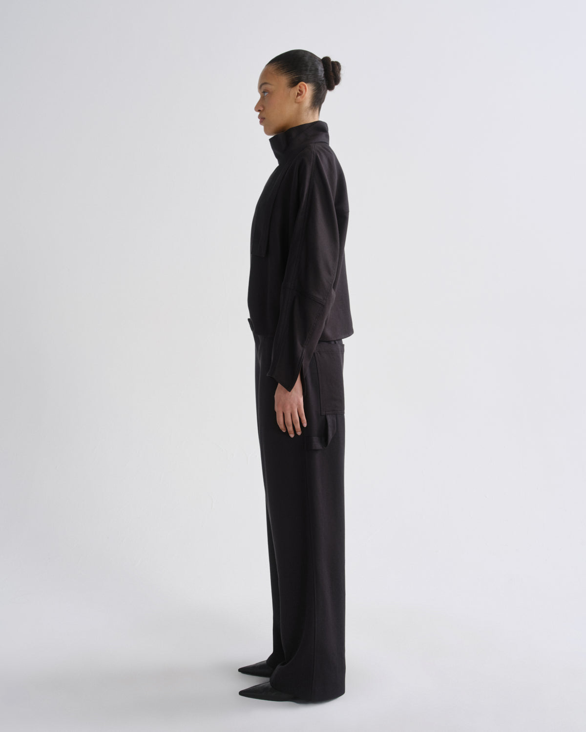 A model stands against a light gray studio background wearing a charcoal long-sleeve mock-neck top with a large bow detail at the collar and matching charcoal wide-leg trousers.