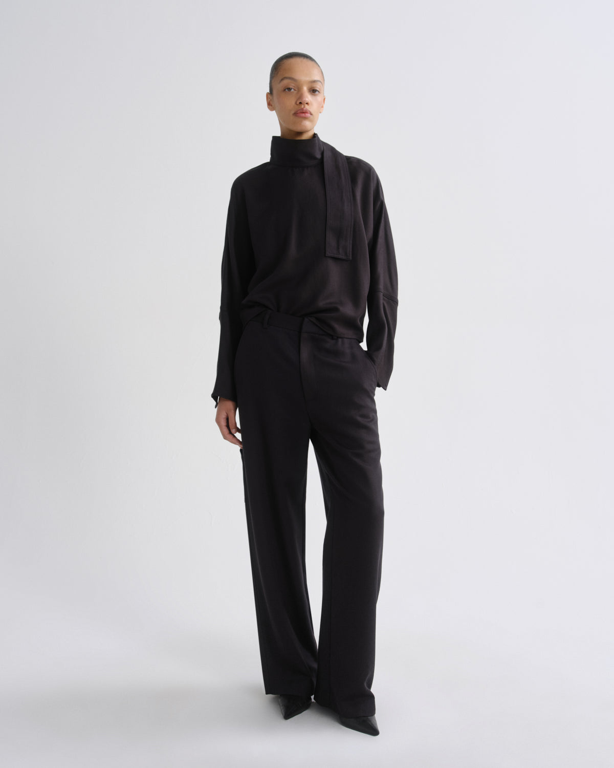 A model stands against a light gray studio background wearing a charcoal long-sleeve mock-neck top with a large bow detail at the collar and matching charcoal wide-leg trousers.