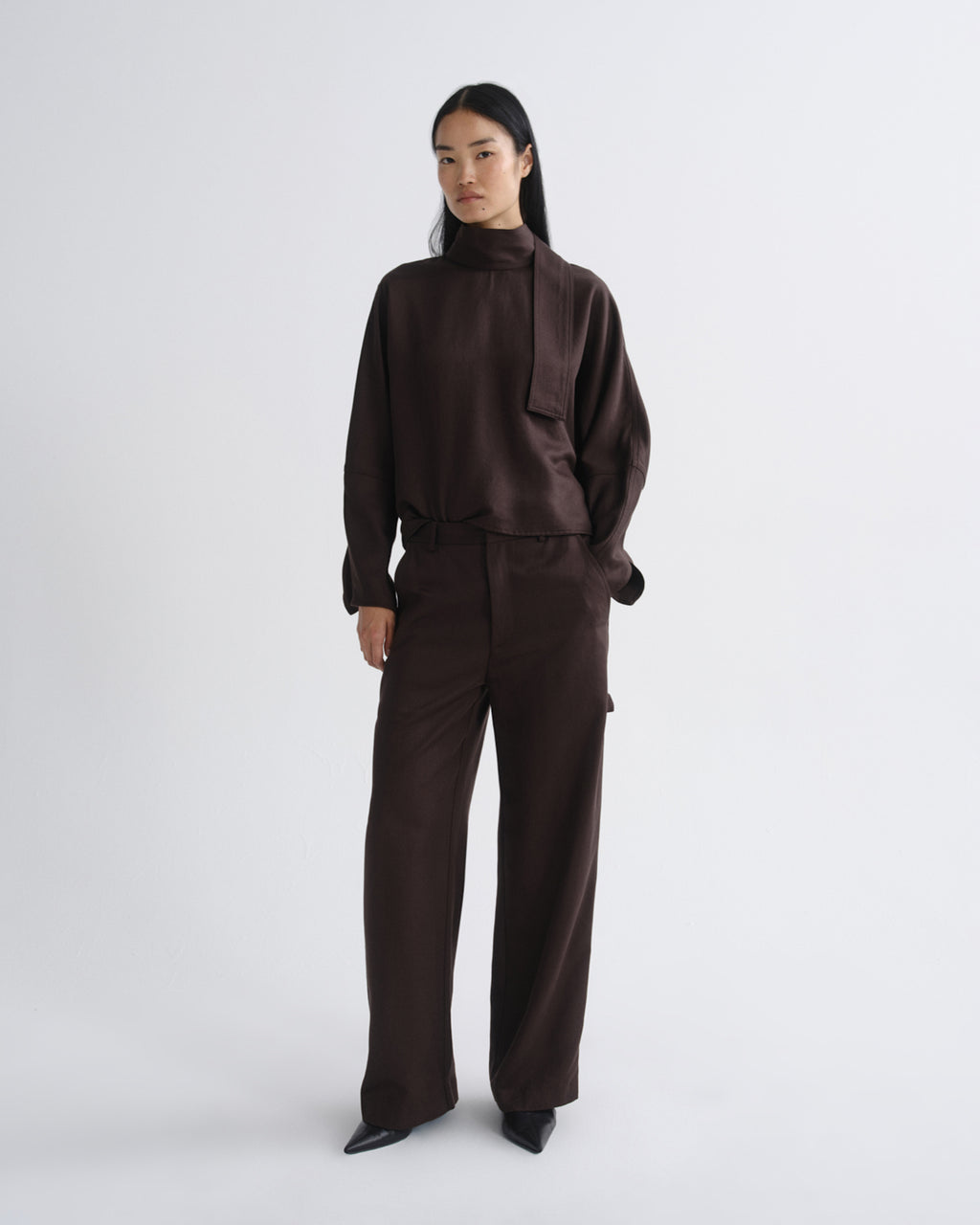 Woman wearing a dark brown long sleeve top with dark brown wide leg pants on a grey background.