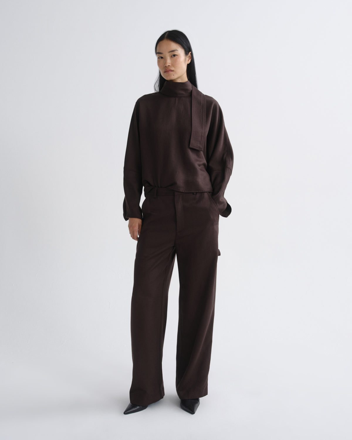 Woman wearing a dark brown long sleeve top with dark brown wide leg pants on a grey background.