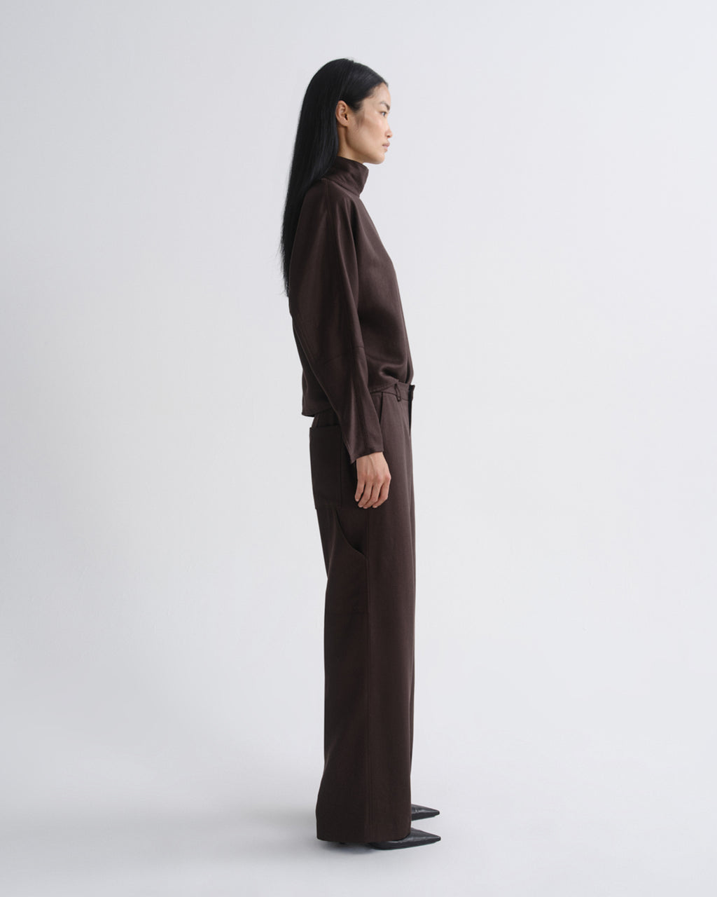 Woman wearing a dark brown long sleeve top with dark brown wide leg pants viewed from the side on a grey background.