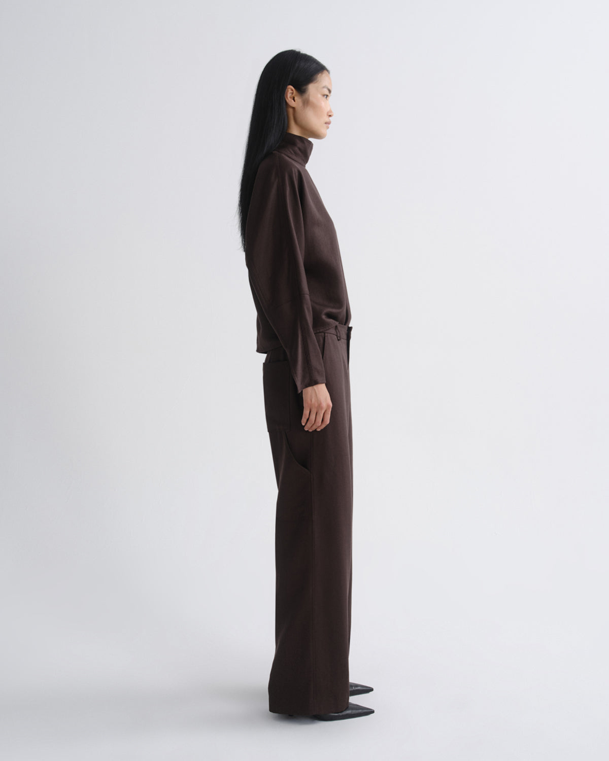 Woman wearing a dark brown long sleeve top with dark brown wide leg pants viewed from the side on a grey background.