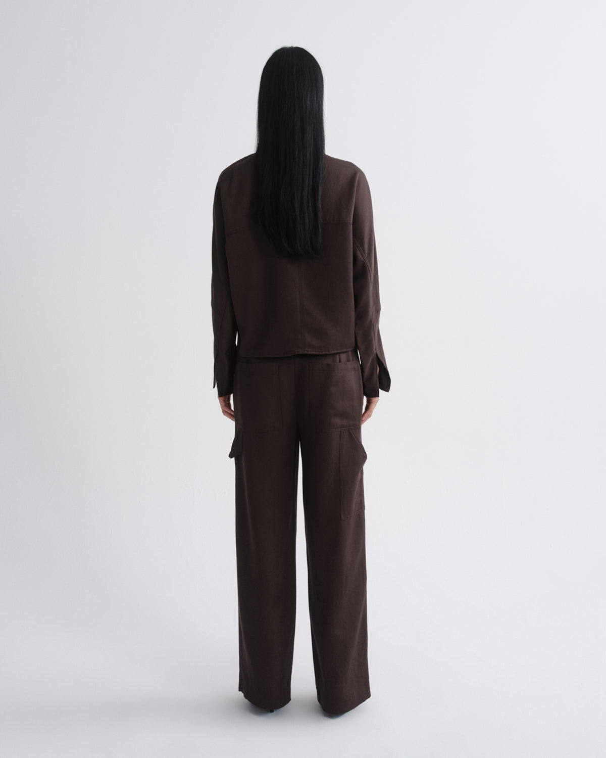Woman wearing a dark brown long sleeve top with dark brown wide leg pants viewed from the back on a grey background.