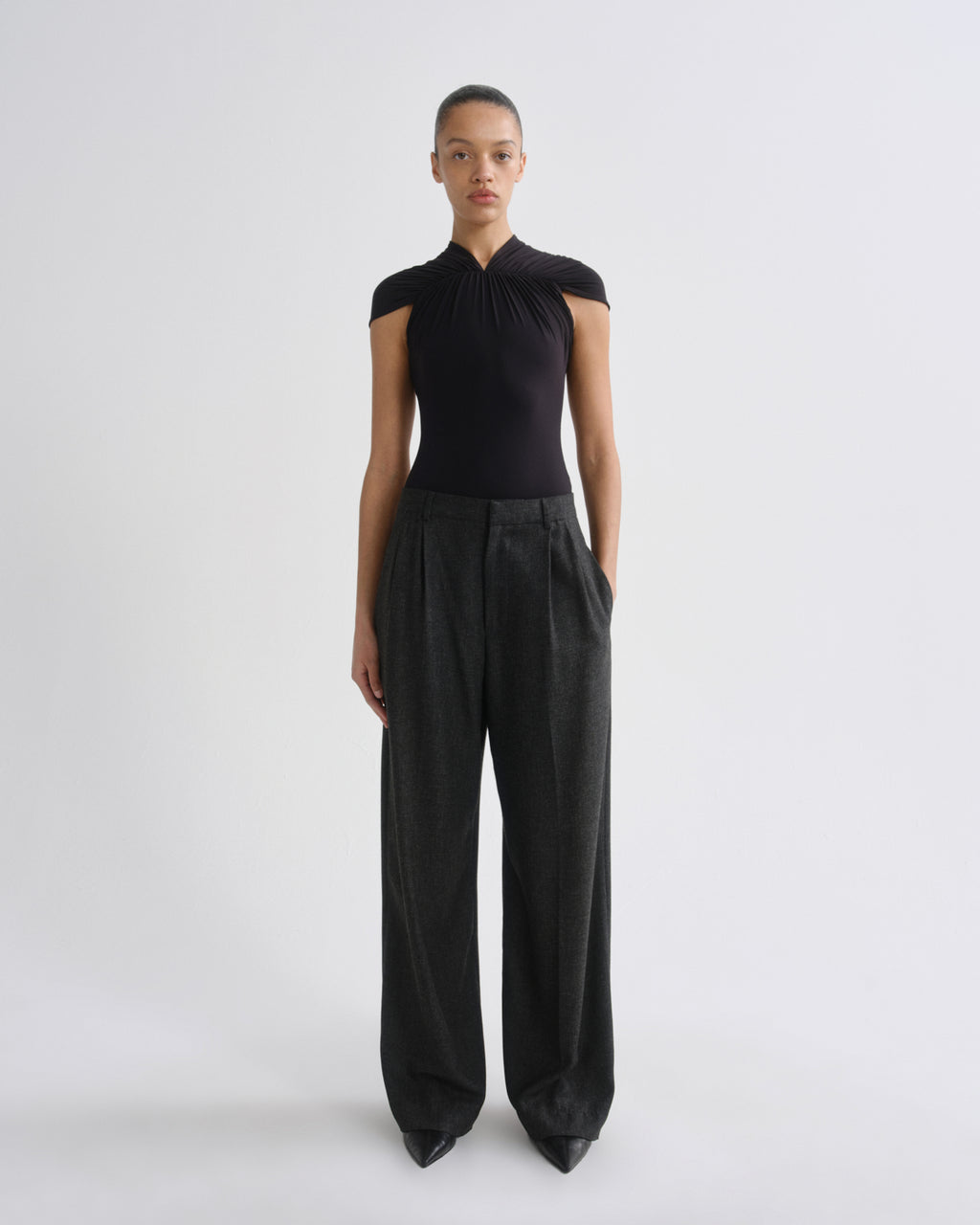 A model stands against a light gray studio background wearing a black sleeveless top with a gathered halter-style neckline and very wide-leg dark charcoal pants.