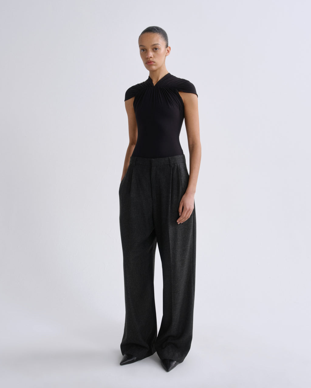 A model stands against a light gray studio background wearing a black sleeveless top with a high gathered neckline and wide-leg charcoal gray trousers.
